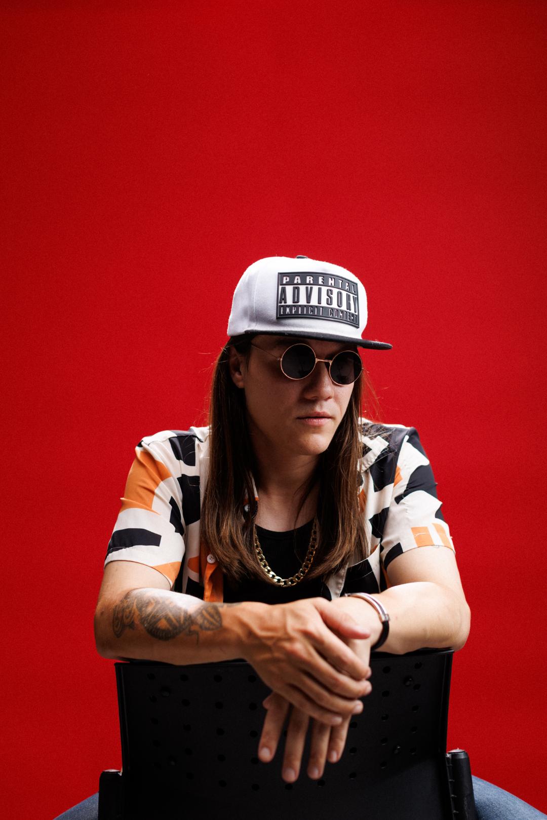 Mon'et press photo. Seated backwards on a chair against a red backdrop, Parental Advisory cap, round sunglasses, gold chain.