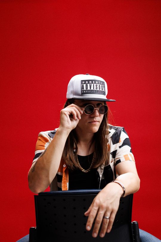 Mon'et lowering his sunglasses, seated on a chair, red backdrop.